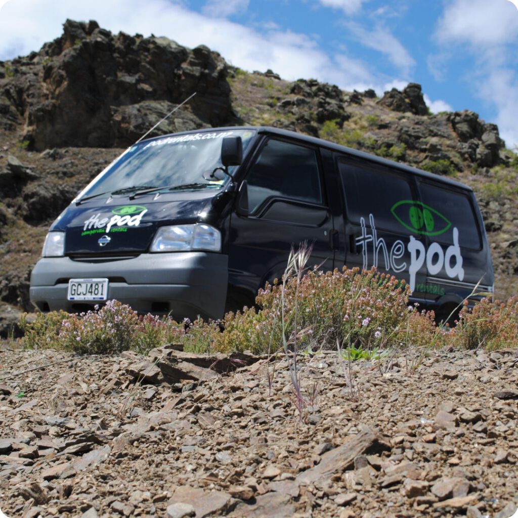 The Pod Camper Van Rental company camper in the terrain it is used for. the photo shows the camper vehicle on a mountain road parked as the independent traveler takes in a local tourism site.